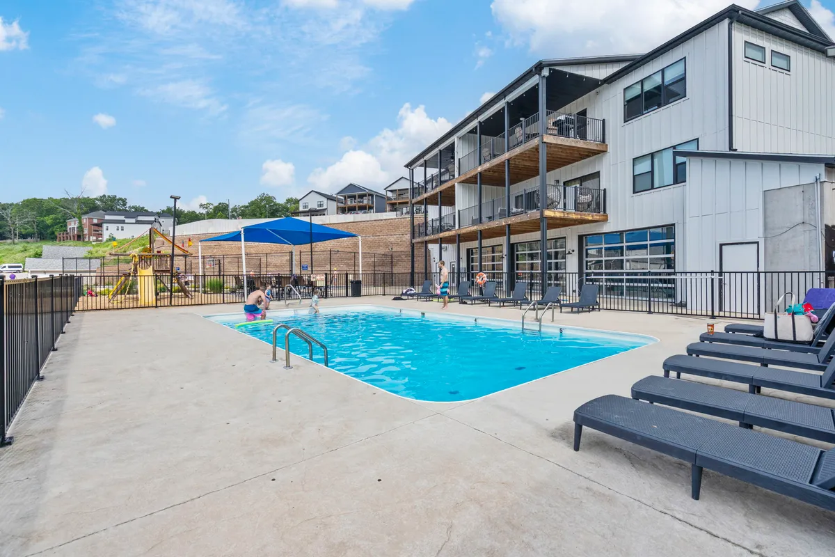This exterior image captures a vibrant communal area featuring a swimming pool, lounge chairs, a play area for children, and balconies of the surrounding building. The space shows people swimming, sunbathing, socializing, and kids playing.