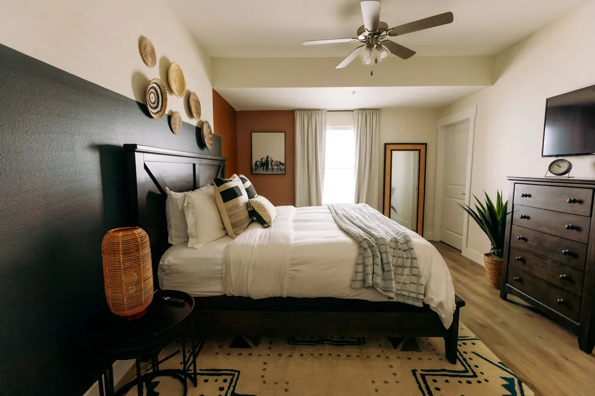 This image displays a serene master bedroom featuring a bed with white linens, woven wall décor, a wooden dresser, a mirror, and an indoor plant. Sunlight gently streams into the room, creating a tranquil and welcoming environment.