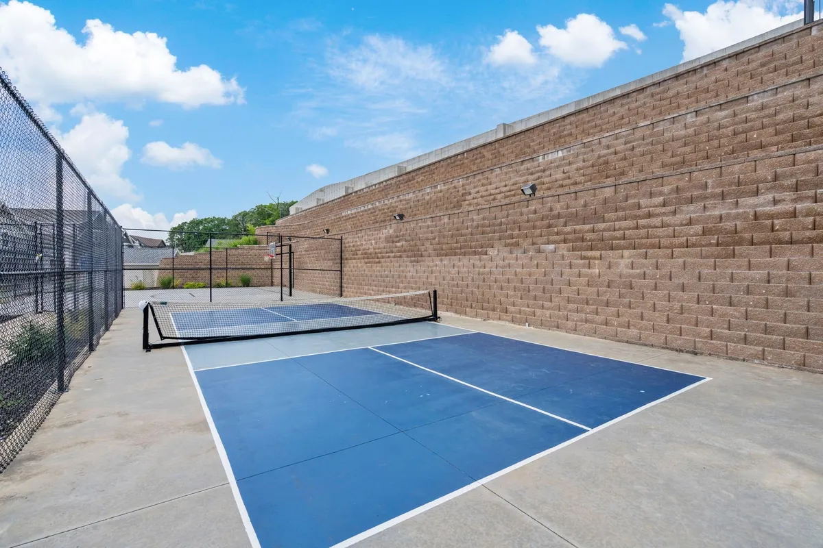 An exterior view of an energetic pickleball court with a chain link fence and brick wall, set against a clear blue sky. The scene captures people playing pickleball and enjoying friendly competition.