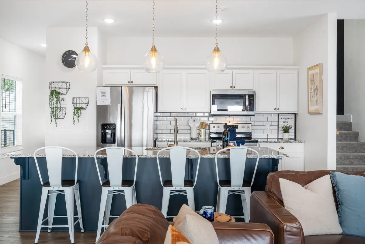 A modern kitchen with a generous island and bar stools, stainless steel appliances, stylish white cabinetry, pendant lighting, and a view into a cozy living area. The design invites cooking and conversation, serving as a stylish and functional hub for meals and gatherings.