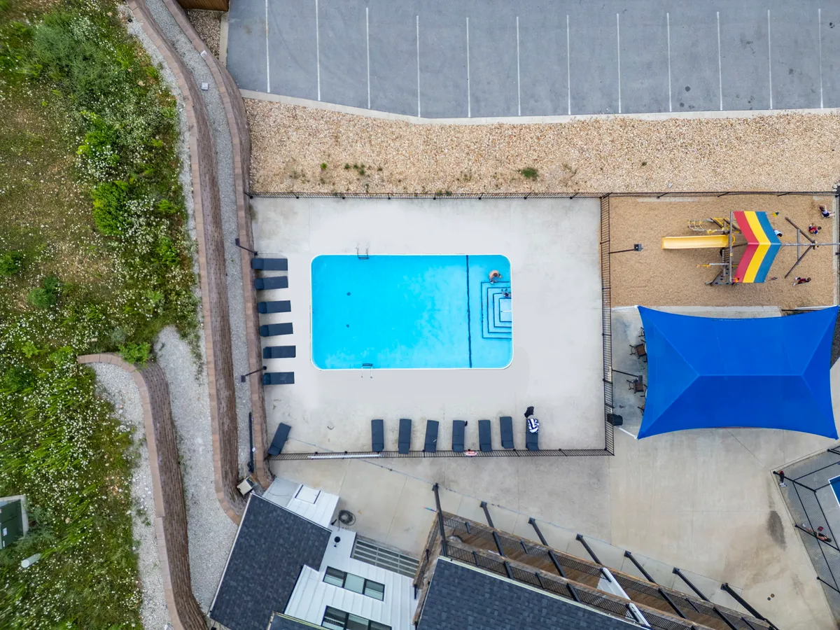 This exterior image showcases a vibrant community swimming pool, complete with lounge chairs and a blue shade structure. Children are actively playing on the nearby playground equipment, while families and friends gather around the pool area. The setting is sunny and lively, reflecting summer fun and communal enjoyment.