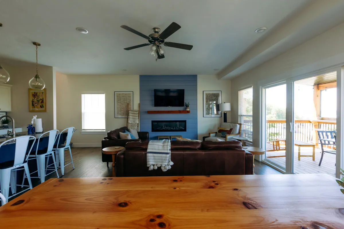 A spacious open-concept living area featuring a comfortable leather sofa, a warm fireplace, a large dining table, and expansive windows that let in abundant natural light. The space seamlessly blends areas for relaxation, dining, and socializing, with the outside deck visible through the windows.