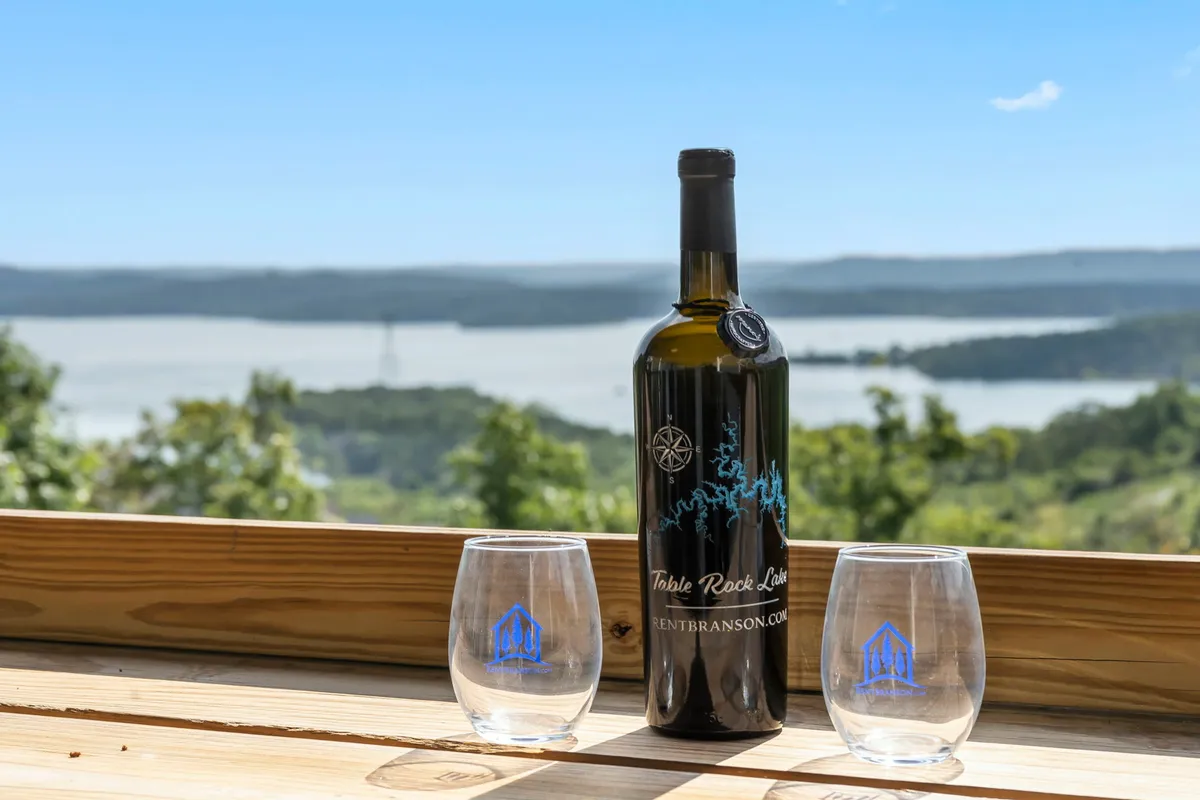 A close-up of a wine bottle and two glasses set on a ledge with a distant lake view and lush greenery in the background. The mood is serene and intimate, ideal for sipping wine at sunset and reflecting on the day’s adventures.