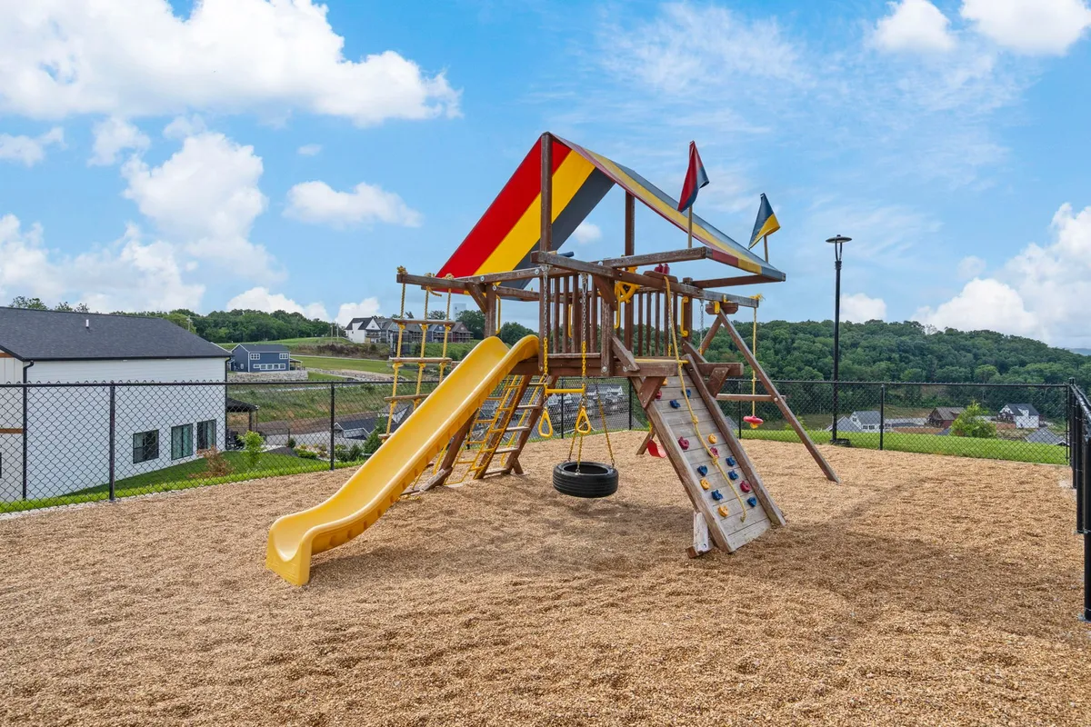 This exterior shot centers on a colorful playground structure featuring a slide, swing, and climbing wall, with children playing and parents supervising nearby. The mood is joyful and lively.