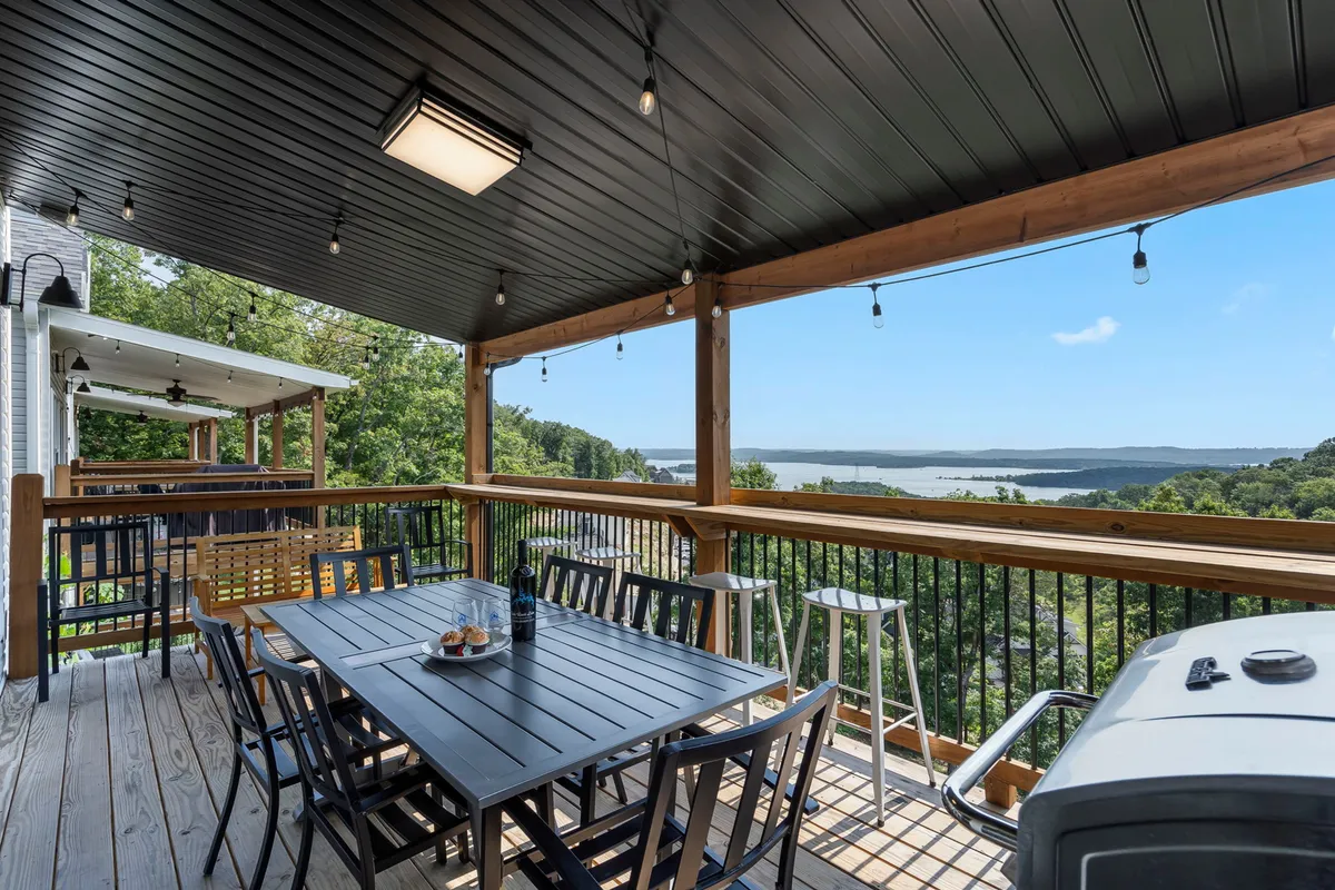 An inviting outdoor dining area on the main-level deck, featuring a dining table, grill, hanging lights, and spectacular lake views. The space is designed for al fresco meals, barbecues, and social gatherings under open skies.