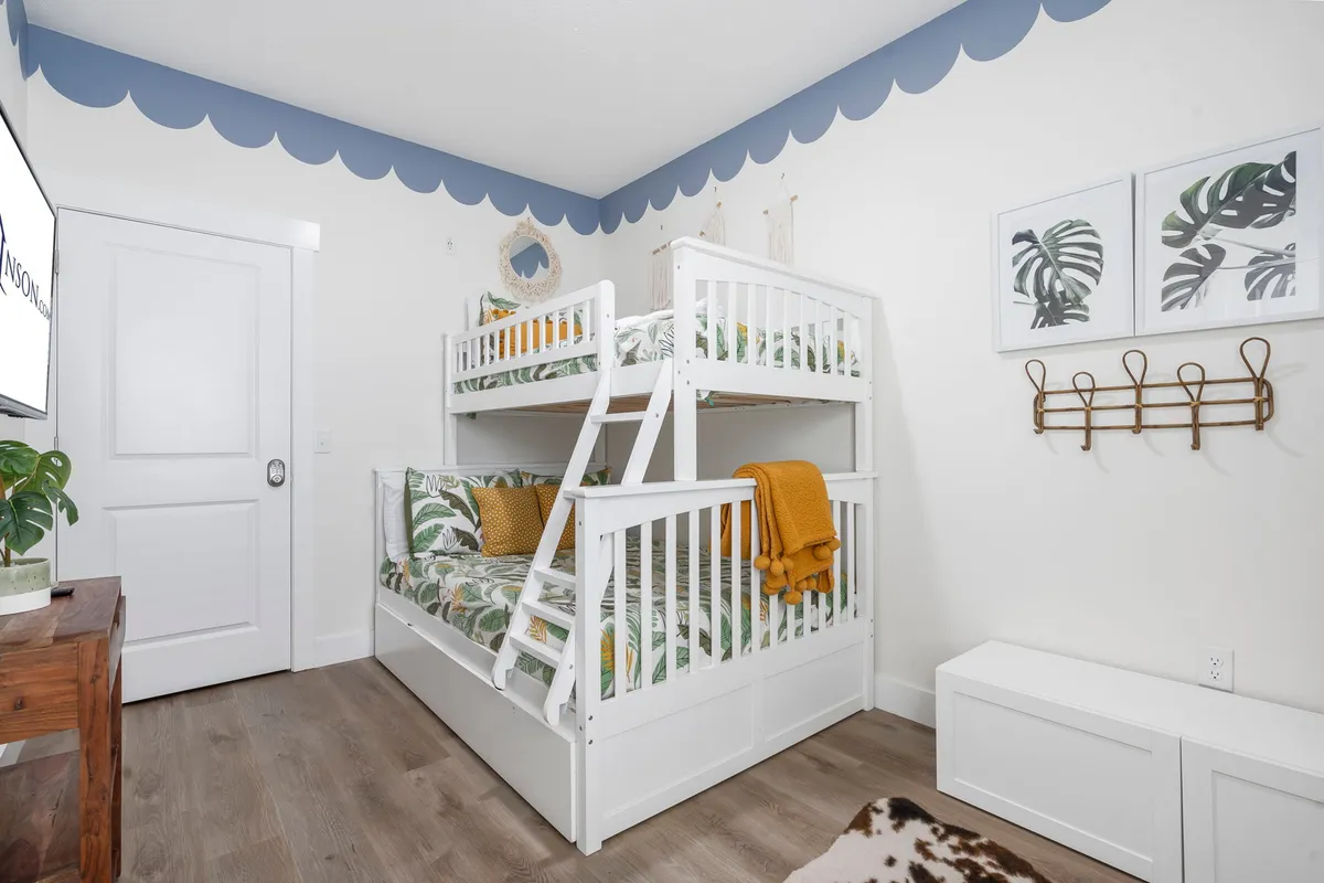 This playful interior shows a bunk room with sturdy bunk beds, whimsical wall art, a plant, and a cheerful yellow throw blanket. The space is cozy, inviting, and clearly designed for children or teens.
