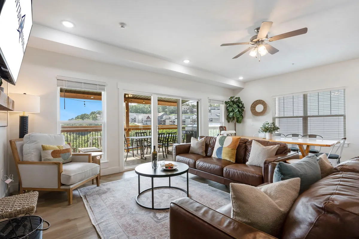 The image features a stylish, modern farmhouse living area with a plush leather sofa and coffee table, bathed in natural light from expansive windows that showcase breathtaking sunset views. The open concept seamlessly connects the cozy lounge space with a spacious dining area, inviting guests to gather, relax, and enjoy meals together while overlooking the lake and mountains beyond.