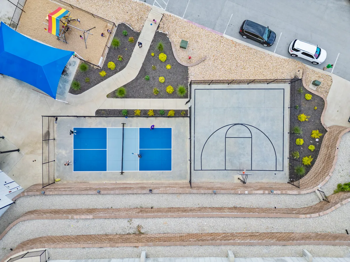 A vibrant exterior scene featuring basketball and tennis courts alongside a playground area, all set within a community environment. The blue shade structure and surrounding green space suggest an active, family-friendly atmosphere perfect for sports and recreation.
